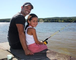 A dad and his young daughter who is holding a fishing rod smile at the camera as they sit by the Hudson River.