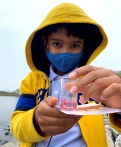 A young boy in a fleece jacket holds out a small jar of water.