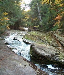 A stream flows between large rocks with Autumn trees in the backgound.