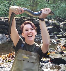 A young woman in waders stands in a stream and joyously holds up an adult eel over her head.