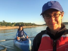 Image of educator Ann Fraoli near a river. A young girl in a kayak is on the river behind her.