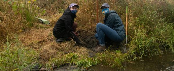 Two women wearing masks plant trees by a stream.