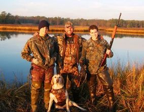 Three men and dog dressed in camouflage pose by the water 