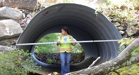 A woman in a neon vest stands in a large concrete culvert measuring its diameter with measuring tape.