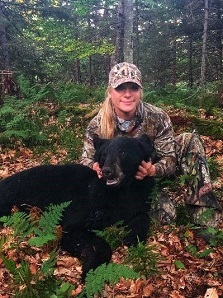 A woman in camouflage poses with a harvested black bear in the woods