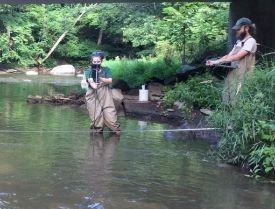 Two people stand in a stream wearing waders to collect water samples.
