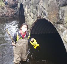 A young woman with a mask stands in a stream next to a stone bridge with a large tape measure.