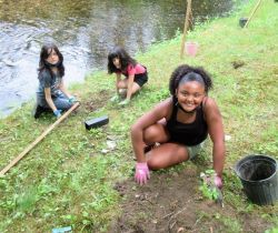 3 young girls plant tree seedlings near a stream.