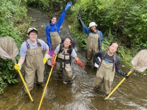 5 young women in waders stand in a stream holding nets.