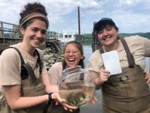 3 young women in waders show a fish bowl of fish from the Hudson River
