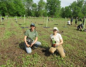 Two women hold pots with tree seedlings in a large field.