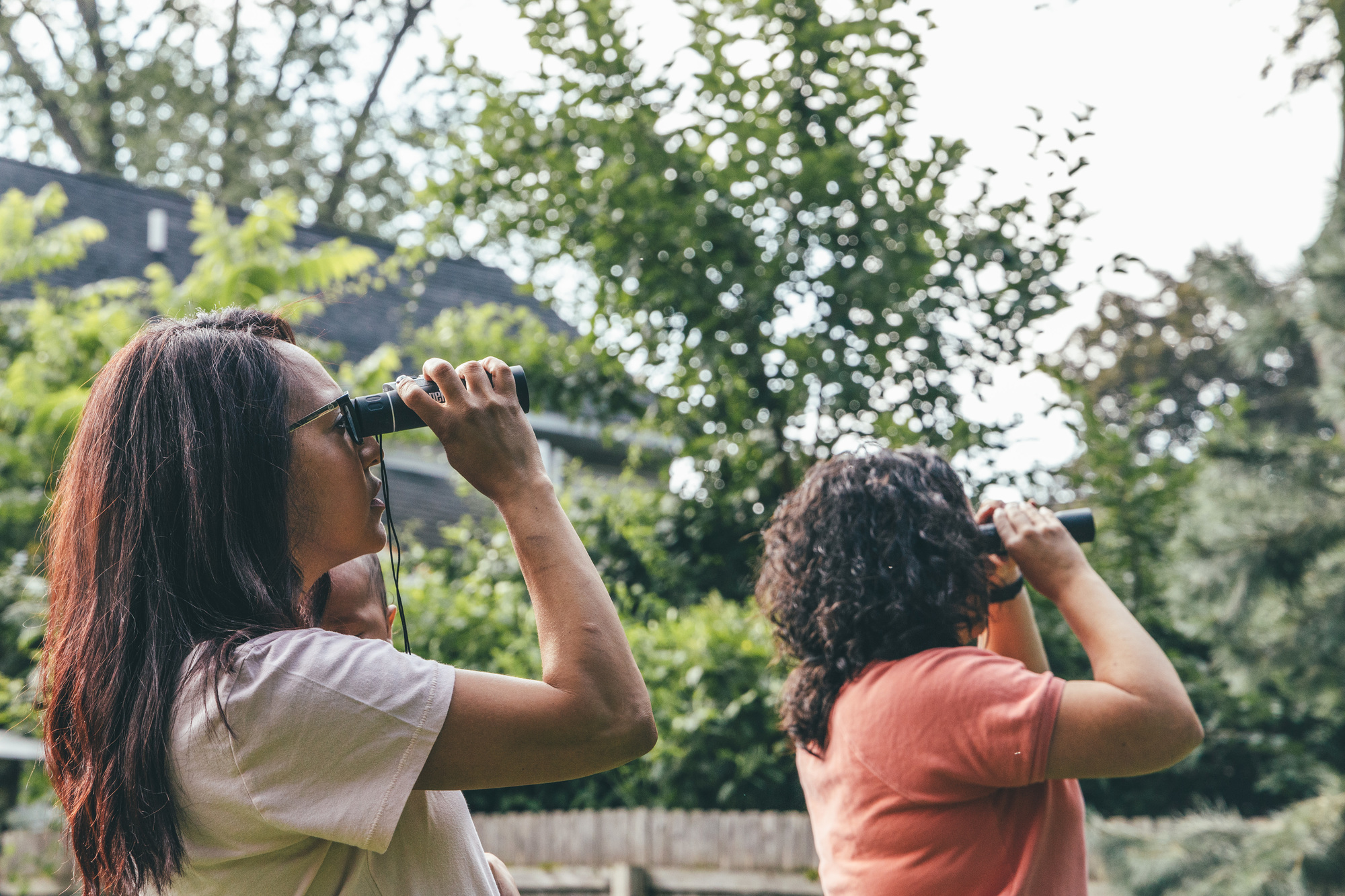Two women standing and using binoculars looking for a bird in the trees