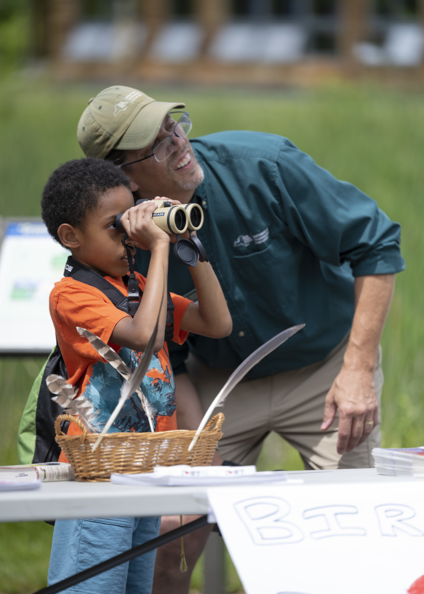 A young boy learning how to use binoculars with a DEC educator looking off in the distance at a bird