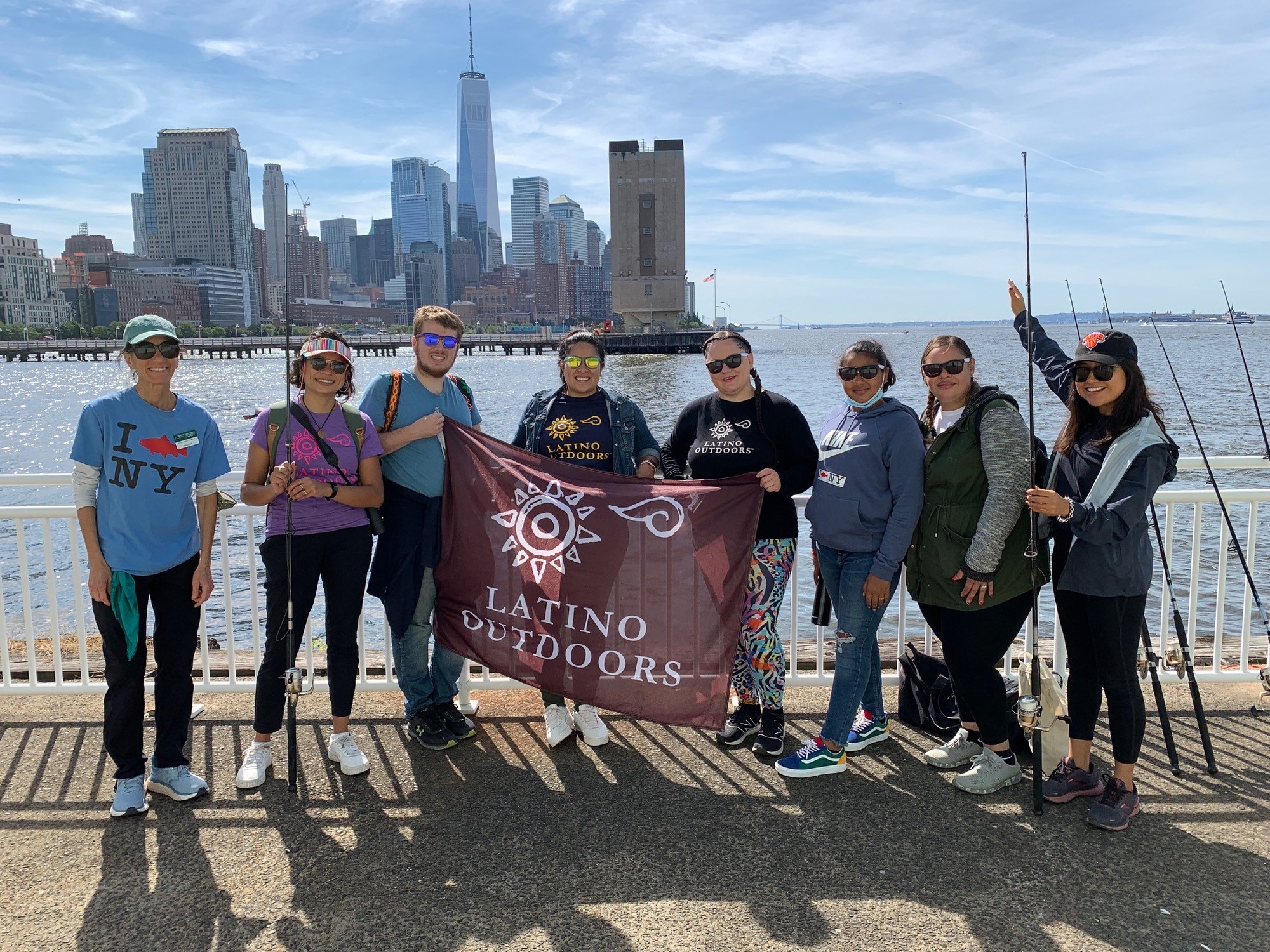 A group of visitors from Latino Outdoors holding a flag that says 'Latino Outdoors' and fishing off a dock in NYC
