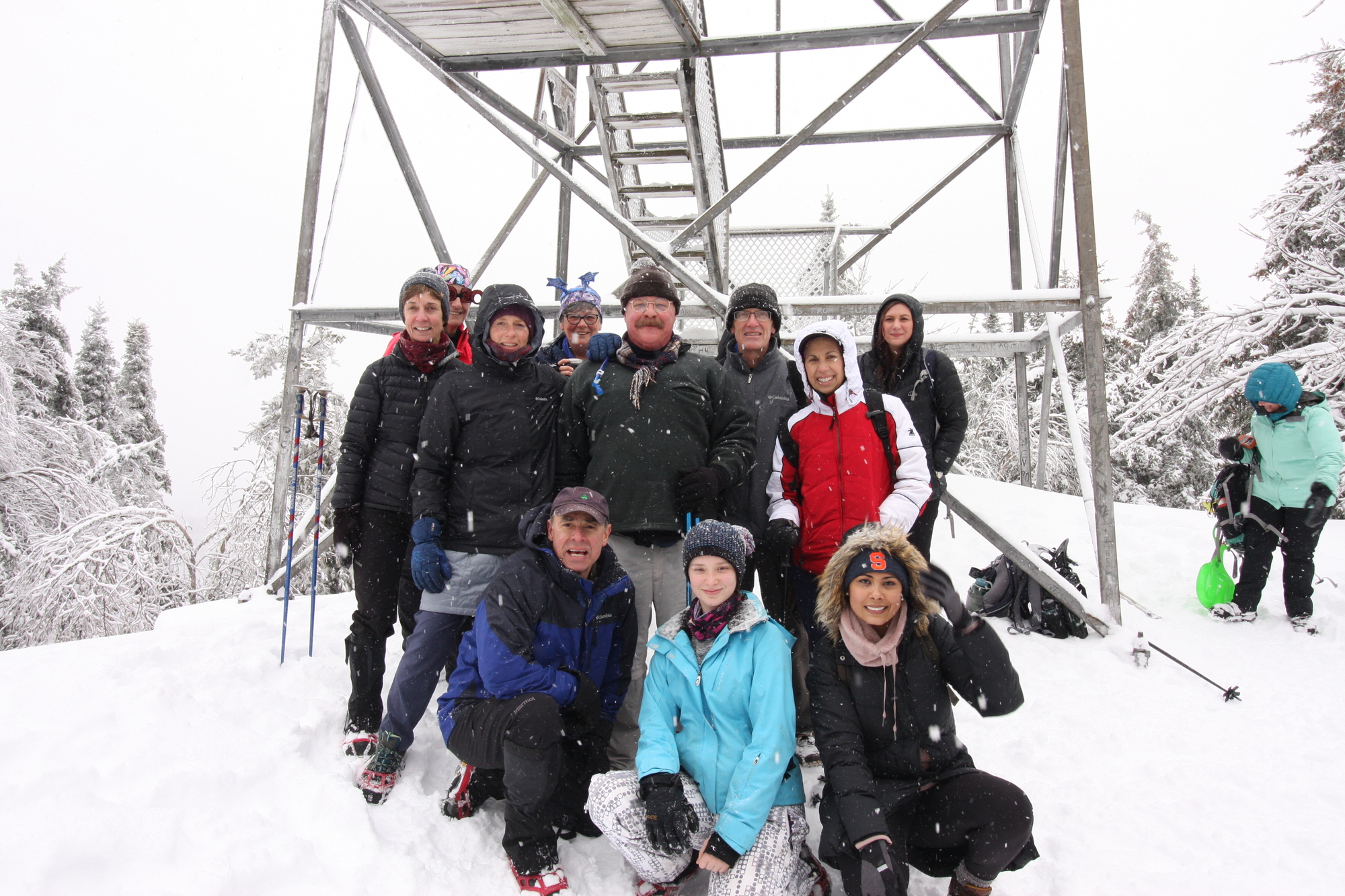 A group of First Day Hikers standing at the base of a fire tower with snow in the background