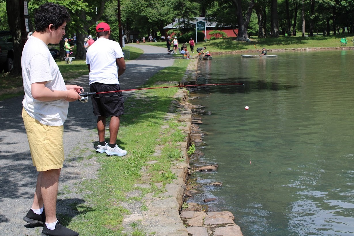 A young person in a light colored t-shirt and yellow shorts holds a fishing pole by the shore of a fishing area.