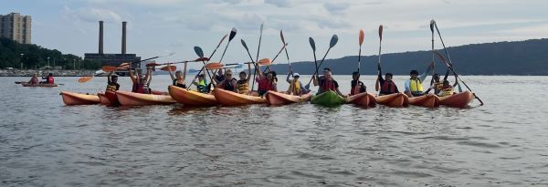 A group of people in canoes on the Hudson River with paddles raised.