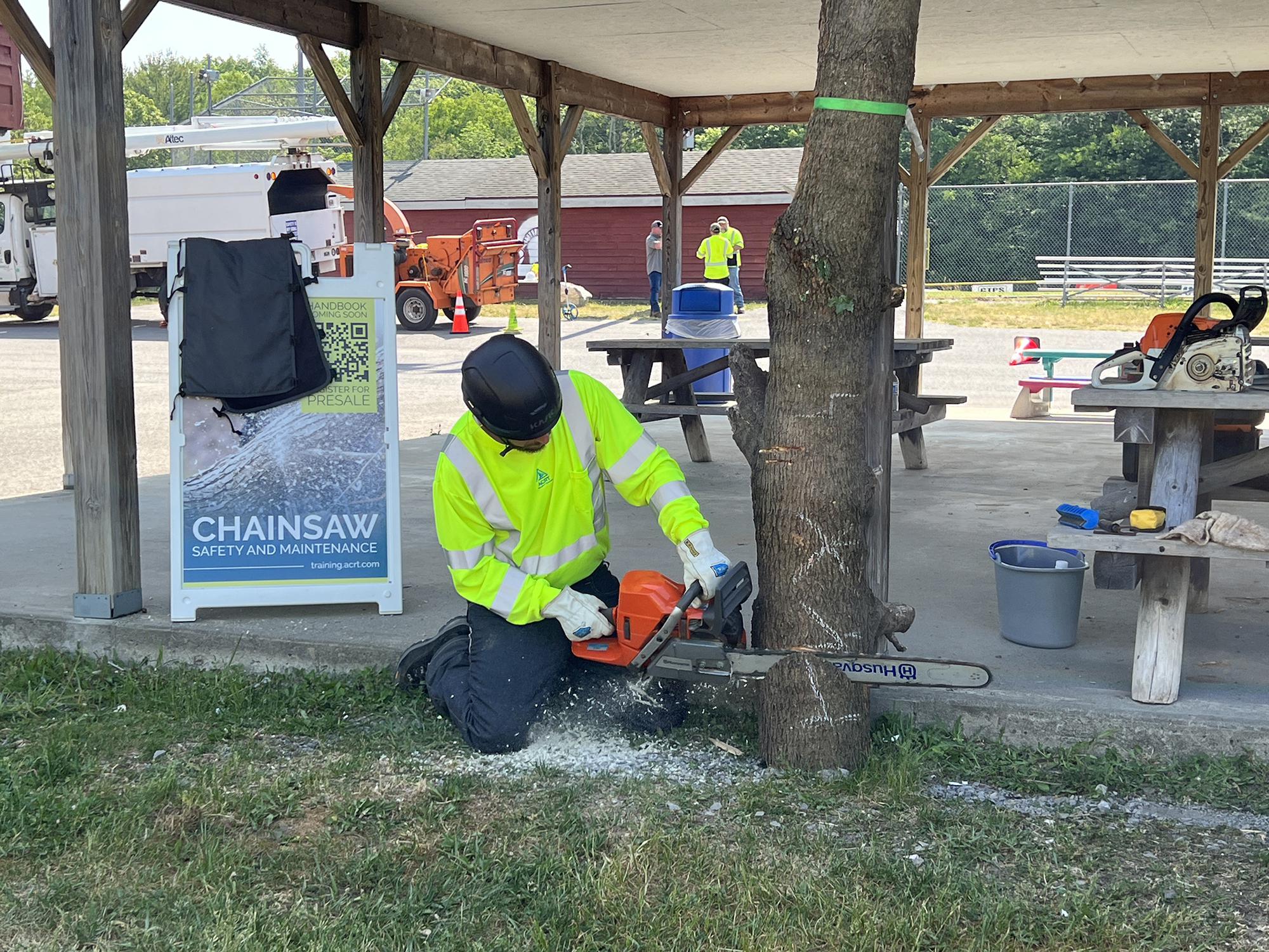 Person in yellow safety vest, gloves, and helmet cutting down hazard tree next to a park pavilion in a chainsaw training course