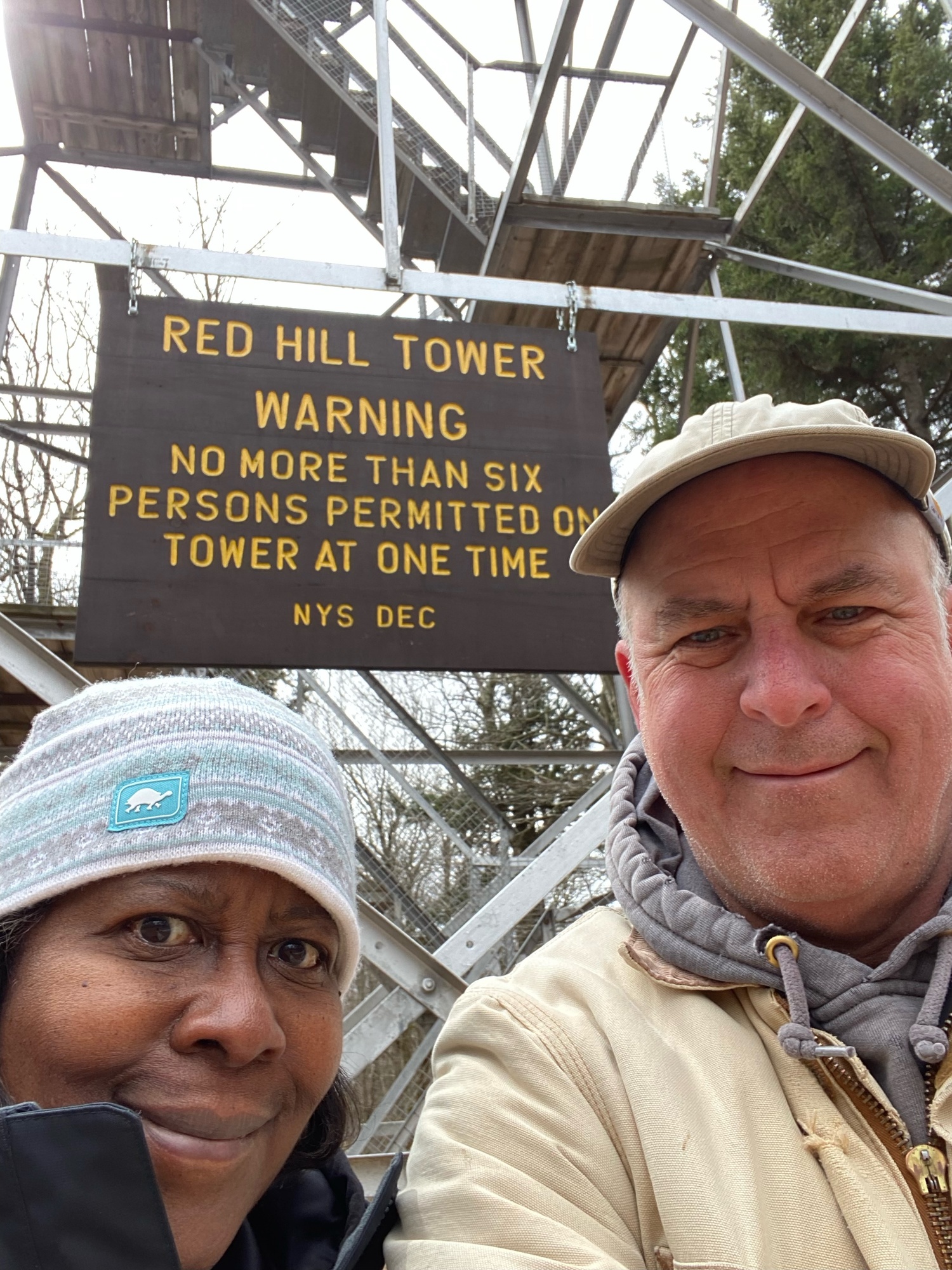 A man and a woman stand in front of the Red Hill Tower sign. The woman is wearing a grey winter hat and the man is wearing a yellow jacket.