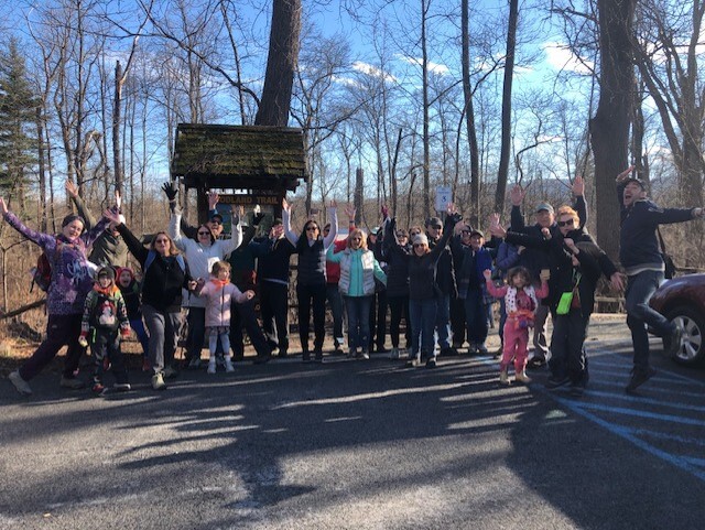 A group of people are standing in front of a sign for the Woodland Trail on a First Day Hike. Many are jumping and waving their arms with excitement. 