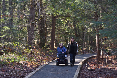 Two people are enjoying a nature trail. A person in a black jacket walks next to a person in a blue jacket, who is sitting in a motorized wheelchair.