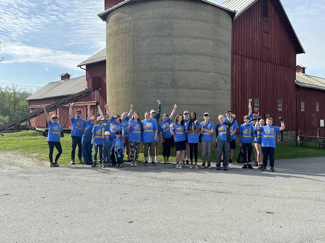A group of people in blue "I Love My Park Day 2024" shirts stand in front of a red barn at Stony Kill Farm Environmental Education Center.
