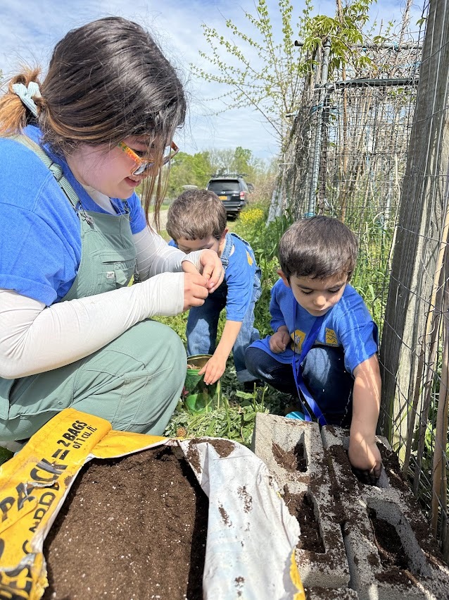 An adult and two children, all wearing blue shirts, plant seeds in upended cinder blocks on a sunny day. 
