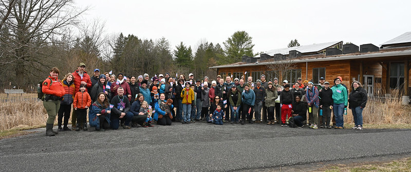 A large group of smiling people pose for a group photo on a cloudy day. Many of them are holding Transgender Pride flags.