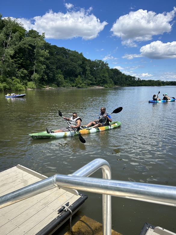 Two people wearing personal flotation devices (PFDs) sit in an open-top kayak and paddle near the dock on a sunny day.