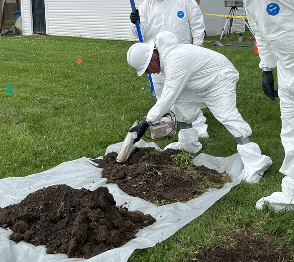 Picture of crew in protective gear testing recently-dug soil samples from a yard. Garage is shown in the background.