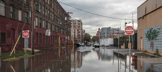A street flooded  with water.  Two signs of "Do not enter" on both sides of the street. A few cars in the background.