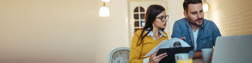woman holding clipboard and man with a cup of coffee looking at a laptop screen