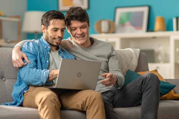 Smiling couple sitting on couch in living room looking at laptop. In background: bright blue walls, book shelf with artwork.