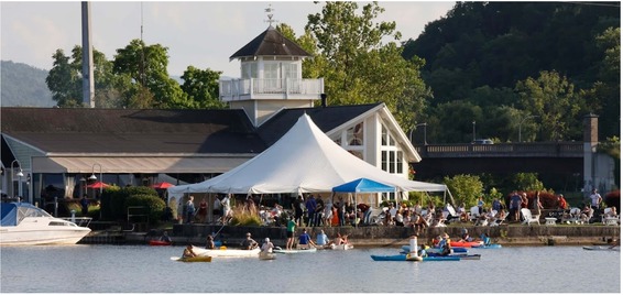 a group of kayakers and paddlers near a large tent with a crowd listening ot a musical performance