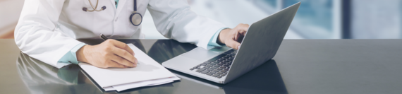 A medical professional working on a laptop and holding a pen with a physical file in front of them.