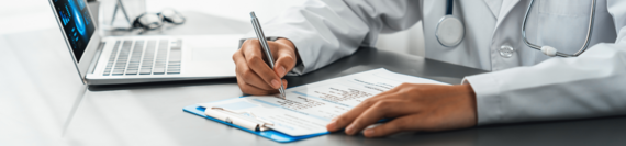 A doctor filling out a paper form while looking at a laptop.