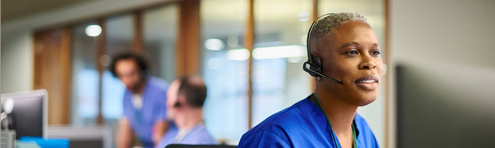 Healthcare worker wearing a headset sits at a workstation, with two other workers visible in the background