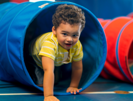 A toddler boy with curly hair crawls through a blue flexible tube.