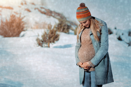 A pregnant women is standing in the snow, bundled up for winter.