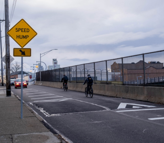 Speed hump with two people riding bikes in the opposite direction of the camera and two people driving cars towards the camera. 
