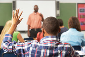Back of student who is raising his hand.  A teacher stands in front of the class facing the students.