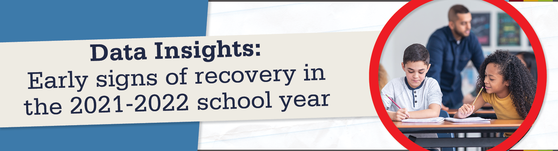 Students at a desk in the classroom. Teacher is in the background. 