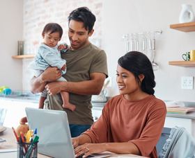 a young woman works on a computer while a young man holds a baby and watches over her shoulder