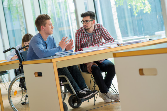 Young man in wheelchair talking with other student in college classroom