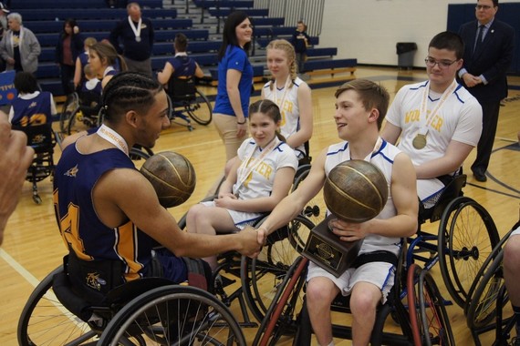 photo of man in wheelchair with basketball smiling at young kids in wheelchair with one boy holding a basketball but all have medals on