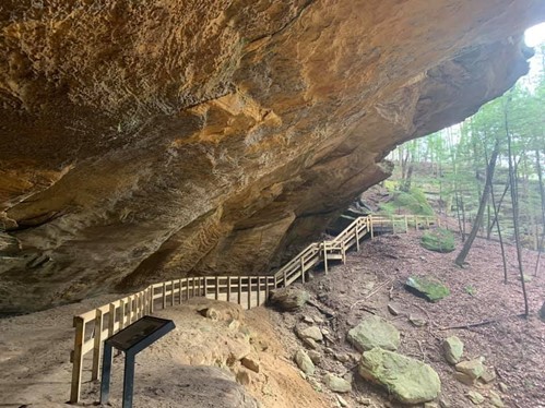 image of giant brown rock with a walk path underneath it and a hand rail with green trees from a distance