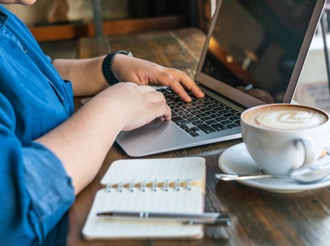 Image of woman's hands on laptop, with a cup of latte and a small notepad and pen next to the laptop