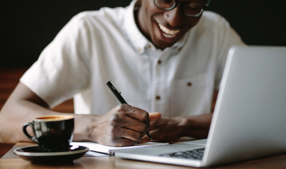 Photo of man smiling, wearing glasses with pen in his right hand looking down at paper with laptop in front of him