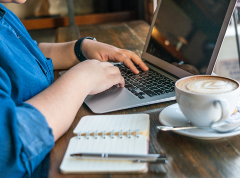 photo of person at desk with hands on laptop keyboard with notebook and coffee