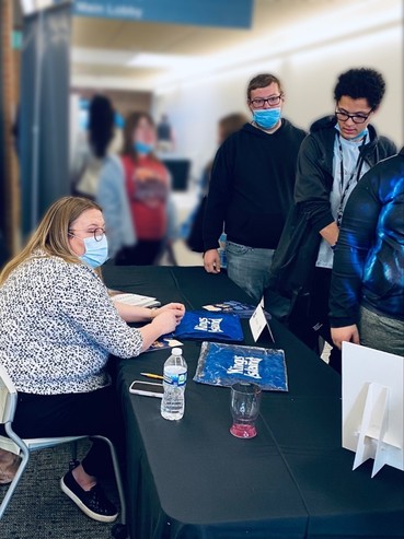 Photo of two students standing in front of a table where a woman is seated
